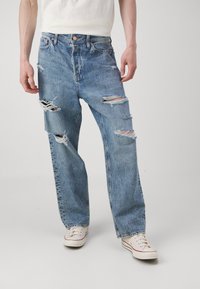 Jean en denim bleu clair avec un effet usé, présentant de grandes déchirures sur les cuisses et les genoux, avec une coupe décontractée et un design classique à cinq poches.