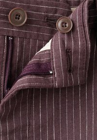 Tela morada con rayas finas, sin cremallera, con dos botones marrones de cuatro agujeros, una cremallera y un forro interior blanco parcialmente visible.