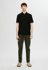 Czarna koszulka polo z krótkim rękawem i stojącym kołnierzykiem, z zamkiem błyskawicznym z przodu, w połączeniu z oliwkowymi spodniami i beżowymi sandałami.