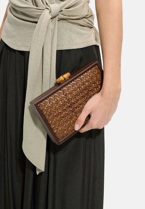 Dłoń trzyma prostokątną plecioną kopertówkę z rattanu z ciemnobrązową ramą, ubraną w beżowy wiązany top i czarną spódnicę na białym tle.