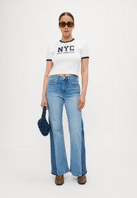 Hvit kortermet t-skjorte med marineblå "NYC" tekst, kombinert med lyseblå fishtail jeans med mørkere sidesømmer og brune firkantede boots.