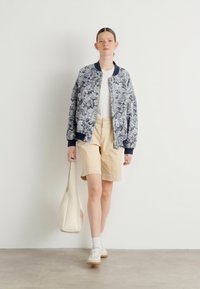 Veste bomber florale avec des accents bleus, t-shirt blanc, shorts beiges et baskets blanches. Porte un sac fourre-tout crème. Fond neutre.
