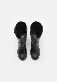 Un par de botas de cuero negro hasta la mitad de la pantorrilla con la punta redonda y tiradores, vistas desde arriba sobre un fondo blanco.