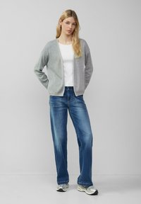 Grauer und hellgrauer Cardigan mit offenem Vorderteil, getragen über einem weißen Hemd und blauen Jeans. Modell steht mit Händen in der Hüfte in einer lässigen Umgebung.