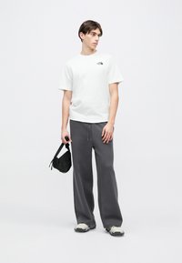 Λευκό κοντομάνικο μπλουζάκι μ ε μικρό μαύρο λογότυπο, σε συνδυασμό με φαρδιά γκρι φόρμα και ανοιχτό γκρι αθλητικά παπούτσια, κρατώντας μια μαύρη τσάντα ώμου.