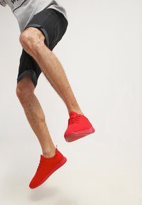 Zapatillas deportivas rojas con parte superior de malla texturizada, puntera redonda y suela de goma plana, combinadas con pantalones cortos negros hasta la rodilla.