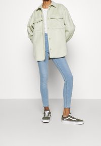 Giacca a camicia oversized verde chiaro con due tasche sul petto, abbinata a jeans skinny blu e sneakers verde oliva con accenti bianchi.
