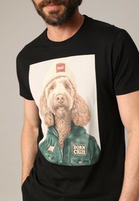 Zwarte T-shirt met een grote afbeelding van een hond met gekruld haar, die een beanie en een groene jas draagt. De tekst luidt "BORN TO CHILL."