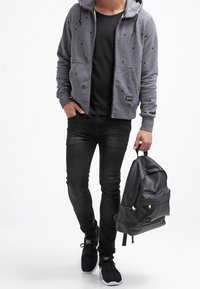 Šedá mikina na zip s hvězdným vzorem, černé tričko, černé skinny džíny a černý strukturální batoh, doplněné o černé tenisky.