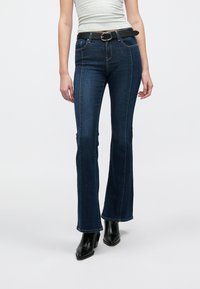 Temno modre zvončaste kavbojke z kontrastnim stranskim šivom, petimi žepi in črnim pasom s srebrno zaponko. Gladek denim material.