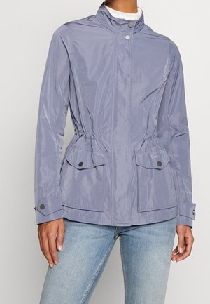 Femme portant une veste zippée violet clair avec poches à boutons-pression et poignets, associée à un jean bleu clair.
