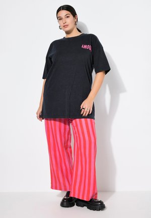 Černé oversized tričko s nápisem "AMOUR" v růžové barvě, doplněné širokými kalhotami s růžovými a červenými pruhy. Masivní černé platformové boty.