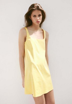 Joven mujer con el pelo largo y castaño, vestida con un vestido amarillo sin mangas, de pie frente a un fondo blanco liso.