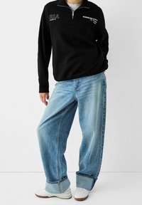 Felpa nera con zip e testo "U.S.A.", abbinata a jeans a gamba larga di un blu chiaro e sneakers bianche con un motivo texture.