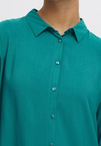 Chemise boutonnée bleu sarcelle avec une texture douce et brossée, dotée d'un col classique et de cinq boutons assortis sur le devant.