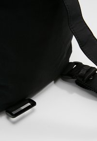 Ecke einer schwarzen Stofftasche mit verstellbarem, gepolstertem Gurt und Kunststoffschnalle auf weißem Hintergrund.