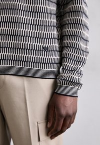 Tumepruun ja kreem patternsiga sviiter soonikkoes, millel on geomeetriline muster. Kergebeezid püksid nähtava tasku jaotusega.