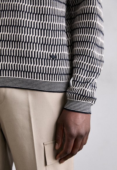 Marineblå og kremfarget mønstrete genser med ribbestrikkede mansjetter, med et geometrisk design. Beige bukser med en synlig lomme.