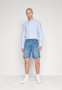 Camisa de botones a rayas azul claro con un bolsillo en el pecho, combinada con pantalones cortos de mezclilla y zapatillas blancas. Tejido suave, corte casual.