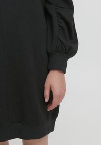 Abito felpa nero con vestibilità ampia, caratterizzato da maniche a sbuffo e polsini a costine. Texture liscia con un aspetto morbido e informale.