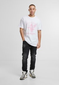 Camiseta blanca de gran tamaño con un gráfico rosa de una figura alada y texto, combinada con jeans negros y zapatillas altas en crema y negro.