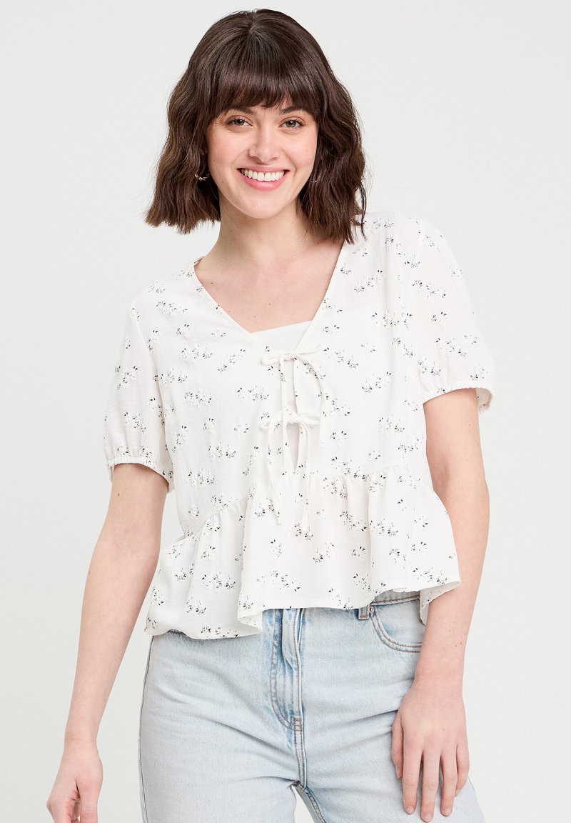 Blouse blanche à peplum avec un décolleté carré et un nœud à l'avant. Présente un motif floral et des manches courtes et bouffantes.