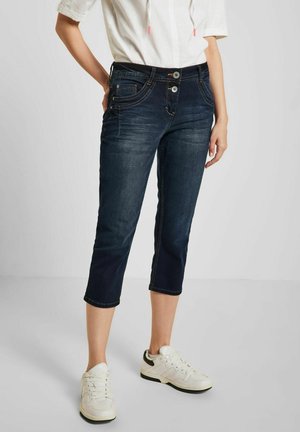 Kvinde iført mørkeblå korte jeans med dobbelte knapper og hvide sneakers, stående med den ene hånd på hoften mod en ensfarvet baggrund.