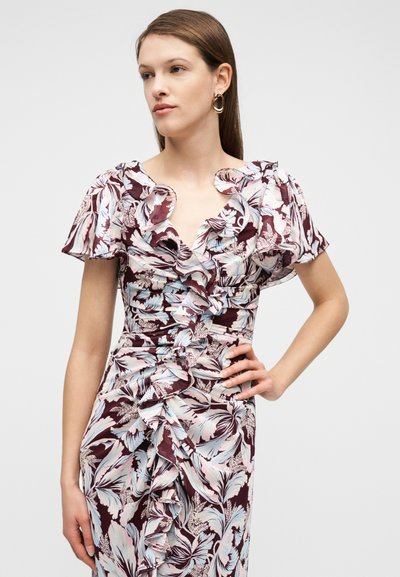 Γυναίκα που φοράει φορεμα με λουλουδένιο βολάν και κοντά μανίκια φτερουγίσματος, στέκεται με το ένα χέρι στη μέση, κοιτάζοντας προς τα πλάγια σε απλό φόντο.