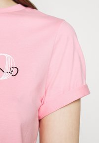 Růžové bavlněné tričko s krátkými rukávy s ohrnutím. Na přední straně je jednoduchý černý grafický design. Hladká textura a volný střih.