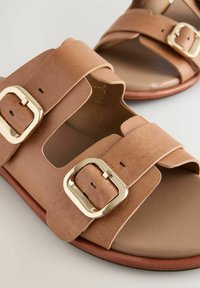 Infradito in pelle color tan con due ampie cinghie, dettagli con fibbie dorate e design aperto sulla punta. La suola è di colore marrone chiaro.