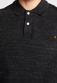 Polo noir en tissu texturé, avec un motif chiné subtil, trois boutons blancs et un logo brodé sur la poitrine.