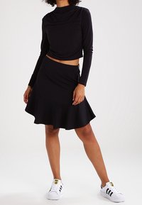 Svart långärmad topp med draperad halsringning, kombinerad med en svart kjol med utsvängd form. Modellen bär vita sneakers med svarta ränder. Mjuk tygtextur.