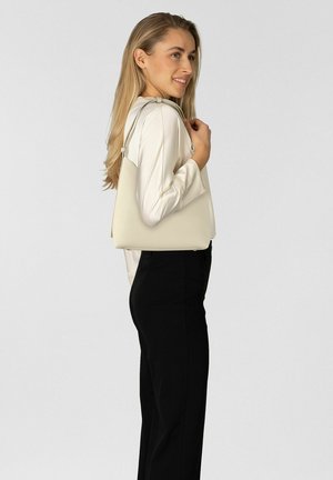 Blonde woman in cream blouse and black pants smiling, carrying a beige leather handbag on her right shoulder against a plain background.
