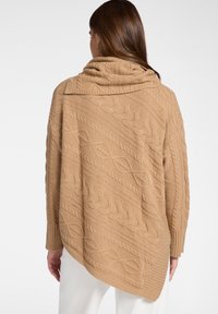 Kobieta ubrana w asymetryczny, beżowy sweter z grubym splotem i dużym, luźnym kołnierzem, ukazana od tyłu na jednolitym tle.