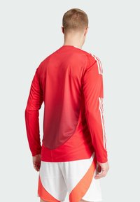 T-shirt de sport rouge à manches longues avec un tissu texturé, présentant des rayures blanches sur les épaules et un ourlet arrière arrondi, associé à un short blanc et orange.