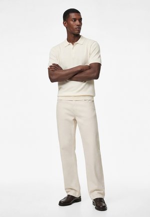 Uomo in piedi con le braccia incrociate, indossa una polo a maniche corte color crema, pantaloni abbinati e scarpe eleganti marrone scuro su sfondo bianco.