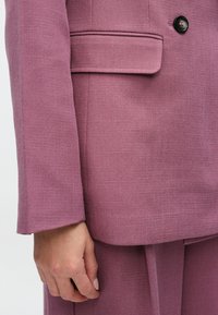 Rosa Blazer- und Hosenanzug mit glatter Textur, Ein-Knopf-Verschluss und Seitentaschendetail, der eine maßgeschneiderte Silhouette zeigt.