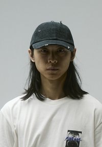 Black denim cap with distressed details and a stitched label reading "PURIFYING ACT OF DEMOLITION." Worn over a plain white T-shirt.