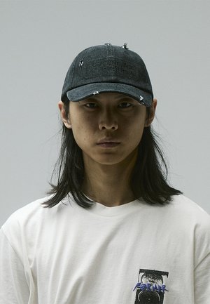 Black denim cap with distressed details and a stitched label reading "PURIFYING ACT OF DEMOLITION." Worn over a plain white T-shirt.