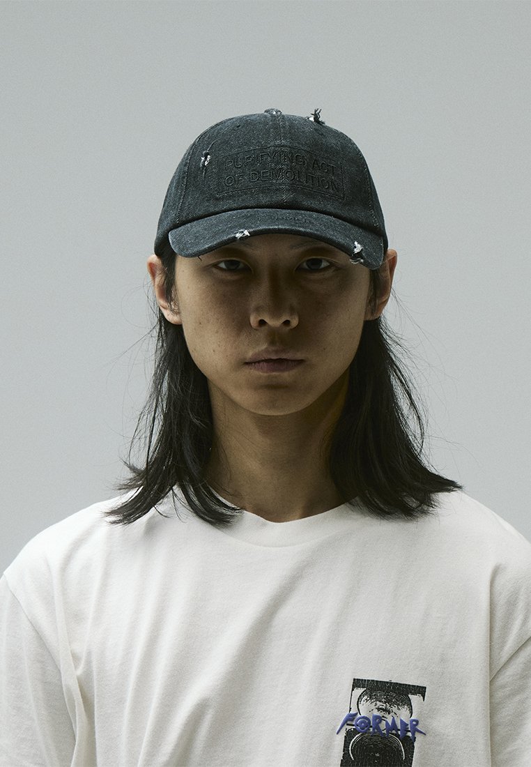 Black denim cap with distressed details and a stitched label reading "PURIFYING ACT OF DEMOLITION." Worn over a plain white T-shirt.