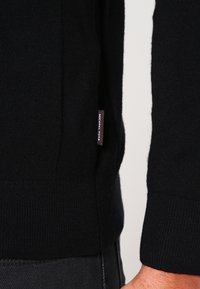 Černý pletený svetr s přiléhavým střihem, žebrovanými manžetami a nášivkou s názvem značky na boku. Textura látky působí měkce a hladce.