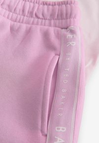 Rosa träningsbyxor med elastisk midja, sidoficka och en kontrasterande vit logoband längs sidan. Slät textur.