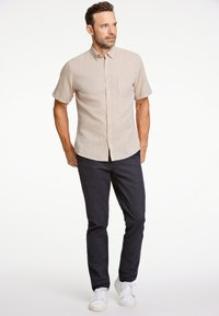 Króka koszula na krótki rękaw w kolorze beżowym z kieszenią, zestawiona z ciemnymi spodniami i białymi trampkami. Koszula wykonana jest z tkaniny o fakturze i ma standardowe zapięcie na guziki.