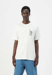 Hvit bomulls t-skjorte med korte ermer. Har et svart grafisk design på brystet og rund hals. Løs passform. Denim jeans.