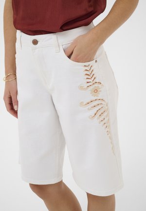 Femme portant un short blanc jusqu'aux genoux avec une broderie beige et un haut couleur rouille, main posée dans la poche, bracelet en or au poignet.