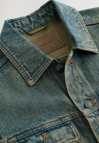 Kék farmerdzseki gallérja és felső része barna varrással, fém gombokkal és egy bézs címkével, amelyen belül "DENIM SUPPLY" felirat olvasható.