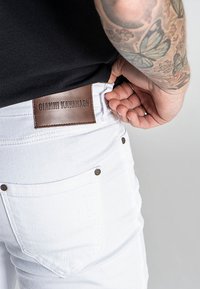 Valkoiset farkut, joissa on nahkainen merkki "GIANNI KAVANAGH" vyötäröllä, hienovaraisella tikkaustyöllä ja metallinapeilla.