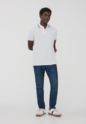 Polo bianco con colletto e maniche a righe nere, abbinato a jeans blu e sneakers bianche con dettagli neri. Design semplice e pulito.