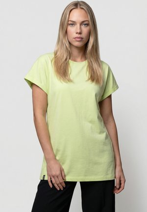 Blonde Frau trägt ein locker sitzendes, hellgrünes T-Shirt mit kurzen Ärmeln und schwarze Hosen, steht vor einem einfarbigen grauen Hintergrund.