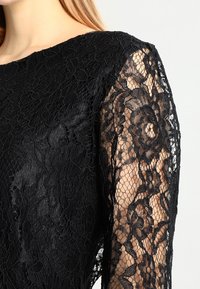 Nära bild av en svart blommig spetsklänning med genomskinliga ärmar som visar huden under, bärs av en person med blont hår.